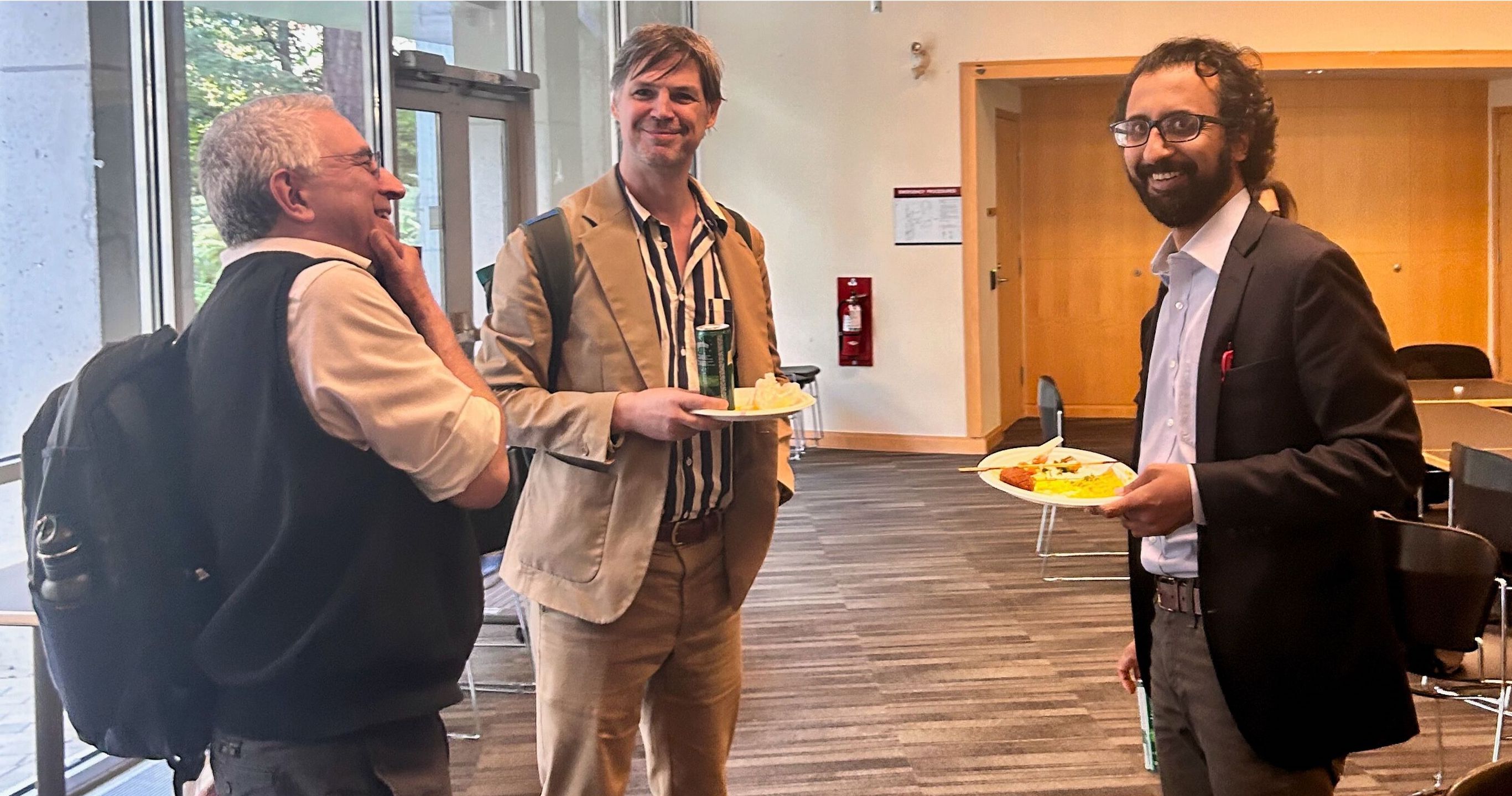 From left to right, John Roosa, Jeffrey Byrne, and Hasan Siddiqui are standing and smiling. They are eating lunch and talking to each other. 