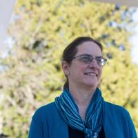 Dr. Barbara Arneil has brown hair, tied in a low ponytail and wears rectangular glasses. She is wearing a teal scarf around her neck and smiles softly off-camera.