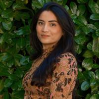 Fiana has black hair and is smiling turned halfway towards the camera. She is standing against a background of leaves. She is wearing a patterned brown top.