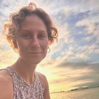 Julia Greening has short, light brown curly hair and is wearing a pink tank top with a floral print. She is looking at the camera, and is positioned in front of the ocean with the sun setting in the background.