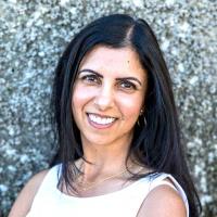 Dr. Renisa Mawani has long black/brown hair and brown eyes. She is wearing a white sleeveless blouse and is smiling towards the camera.