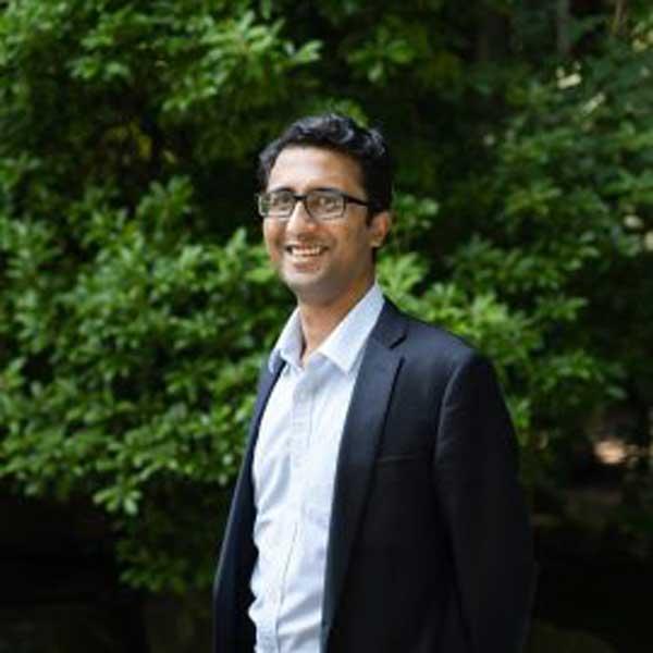 Hasan Siddiqui has short dark hair, glasses, and is wearing a white collared shirt and dark blazer. He is smiling towards the camera, and standing outside in front of green foliage.