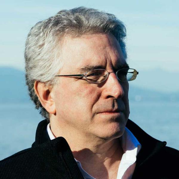 Dr. John Roosa has short grey hair, and is wearing glasses a white shirt and a black zip-up jacket. He is looking to the side and behind him is the ocean and mountains.