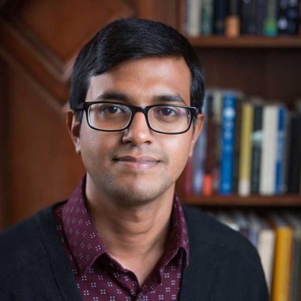 Dr. Nazmul Sultan has short dark hair and is wearing glasses, a dark red collared shirt and navy cardigan, standing in front of a book shelf, smiling towards the camera